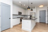 This single-floor unit at 545–555 North Ave NE in Atlanta's Old Fourth Ward neighborhood offers a clean, modern aesthetic throughout its open-concept layout. The kitchen serves as a focal point, featuring flat-front white cabinetry extending to the ceiling, dark stone countertops, and a geometric hexagonal tile backsplash. A central island with a matte black faucet and integrated sink provides additional prep space, while stainless steel appliances—including a gas range, built-in microwave, and French door refrigerator—complete the well-equipped space. Stylish pendant lights with warm-toned interiors hang above the island, adding a contemporary design element. Light hardwood-style flooring runs throughout the main living and kitchen areas, contributing to a bright, cohesive feel. The living area benefits from natural light through large windows with blinds and views of greenery outside. The bedroom features soft carpeting, a ceiling fan, and a generously sized window. The bathroom includes a glass-enclosed walk-in shower with tile surround and hardwood-style flooring, with direct access to a carpeted walk-in closet. The overall ambiance is modern, clean, and well-appointed.