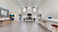 This single-floor community clubhouse offers a generously sized, open-plan layout designed for both social gatherings and everyday convenience. The centerpiece of the main lounge is a dramatic floor-to-ceiling stacked stone fireplace, topped with a flat-screen TV and a rustic wooden mantel, flanked by French doors that provide views of an outdoor pool area. Soaring ceilings, accented with crown molding and dual ceiling fans, amplify the sense of openness, while tiered windows draw in abundant natural light. Light-toned luxury vinyl plank flooring runs throughout, complementing the neutral warm-gray and white color palette.

The lounge area is furnished with leather armchairs, a plush sofa with accent pillows, and layered area rugs, creating a comfortable and inviting atmosphere. A white shiplap bar counter with gold-accented stools serves as a casual gathering spot. Adjacent to the main area, a reception desk and a relaxed seating nook provide a welcoming entry point. A standout fully equipped kitchen features bold navy-blue cabinetry with gold hardware, mosaic tile backsplash, stainless steel appliances, and generous counter space, making it ideal for catered events or resident use.