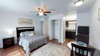 This efficiently designed studio layout makes smart use of its single-floor footprint, offering a cohesive open-plan living environment. The main living area accommodates a full-sized bed and a small dining set, unified by warm wood-style flooring and a patterned area rug that adds visual interest underfoot. A ceiling fan with an integrated light fixture centers the room, providing both air circulation and ambient lighting.

One of the studio's notable functional features is its dual closet arrangement—two side-by-side reach-in closets positioned along an interior wall, offering generous storage capacity rarely found in compact studios. The open closet reveals a full-width hanging rod, maximizing organization potential.

Transitioning seamlessly from the living area, a dedicated kitchenette is tucked into an alcove along the back wall. It features dark cabinetry, a sink, and counter space, providing essential cooking and prep functionality. The white walls and light ceiling keep the space feeling open and bright. Overall, the studio presents a clean, practical layout suited for individuals seeking a streamlined, low-maintenance living space.