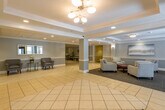 This single-floor common area presents a polished and well-maintained interior designed to serve as a welcoming hub for residents and visitors alike. The space is defined by wide, light-filled corridors featuring large-format cream tile flooring laid in a diagonal pattern, which adds a sense of depth and spaciousness throughout. Crown molding and recessed lighting provide a refined architectural character, while soft blue-gray wall tones contribute to the calm, neutral ambiance.

A central lounge area anchors the space, furnished with a set of upholstered armchairs arranged around a rich wood coffee table atop patterned carpeting — creating a comfortable and inviting gathering spot. Additional seating nooks are positioned along the corridors, offering quiet spots for casual conversation. Decorative touches such as framed artwork, a landscape canvas, metallic wall sculptures, and lush potted palms bring warmth and life to the environment.

A service counter with a bulletin board and reading materials provides a functional focal point near the main circulation path. Elevator banks are conveniently located and clearly marked, accompanied by framed artwork and greenery. Hallways extend from the central areas, leading to further building amenities and residences. Overall, the space balances functionality with an understated elegance suited to a well-kept residential building.