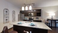 Located at 6634 Riverside Drive in Dublin, Ohio, this single-story space showcases a well-appointed kitchen as a central highlight. The kitchen is designed with rich, dark espresso-toned cabinetry that extends to the upper walls, providing generous storage throughout. A large granite-topped island serves as the focal point, offering seating for casual dining and an integrated sink for added convenience. Three pendant lights hang above the island in a linear track configuration, casting warm, focused illumination over the workspace.

Stainless steel appliances, including a refrigerator, microwave, and range, complement the dark cabinetry and contribute to the kitchen's cohesive, modern aesthetic. The light-toned granite countertops provide a striking contrast against the darker elements, creating a balanced and visually appealing design. A built-in wine rack adds a refined touch to the upper cabinetry. The overall ambiance of the kitchen is polished and functional, blending contemporary finishes with practical design elements suited for everyday living and entertaining.