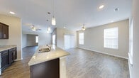 This single-floor home offers a thoughtfully designed open-concept layout that seamlessly connects the kitchen, dining, and living areas into one cohesive, flowing space. The kitchen serves as a central hub, featuring a granite countertop island with an undermount sink, dark cabinetry, and stylish pendant lighting that adds a modern touch. Recessed lighting throughout complements the natural light streaming in from multiple windows dressed with vertical blinds. The expansive living area is finished with smooth, light-toned wood-look flooring that runs throughout, creating a clean and consistent aesthetic. Ceiling fans in both the main living space and an adjacent room ensure year-round comfort. The neutral wall tones and white ceilings contribute to a bright, airy ambiance, making the space feel open and welcoming. The open sightlines from the kitchen island to the far end of the living area highlight the generous scale of this home. Every detail, from the clean trim work to the recessed lighting placement, reflects a contemporary and functional design well-suited for everyday living and entertaining alike.