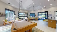Located at 2450 Lake Debra Dr in Orlando, FL, this single-floor community clubhouse is designed to serve as a vibrant social hub. The space is defined by soaring ceilings, large-format white tile flooring, and an abundance of natural light that flows in through oversized windows and a striking glass-panel garage-style door that opens the interior to the outdoors.

The main gathering area centers around a solid wood billiards table set atop a colorful area rug, creating a natural focal point for recreation. Recessed lighting throughout the tray ceiling complements the ample daylight, while the open layout allows for seamless flow between activity zones.

Adjacent to the game area is a comfortable lounge space furnished with teak-framed sofas and chairs accented by vibrant lime green and blue cushions. Large windows dressed with navy curtains frame lush outdoor greenery and pool views, reinforcing the connection to the surrounding landscape.

A functional kitchenette runs along one side of the space, equipped with granite countertops, warm wood cabinetry, and a stainless steel sink — well-suited for catering events or community gatherings. A glass-paneled door provides direct access to the outdoor amenity areas.

Overall, the clubhouse delivers a bright, welcoming atmosphere that balances recreational functionality with comfortable social spaces.
