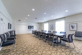 Located at 434 Berkshire Dr in London, Ontario, this well-appointed multi-purpose room offers a refined and functional space suitable for meetings, gatherings, and events. The room is thoughtfully designed with classic architectural details, including white wainscoting panels that line the walls, recessed lighting, and a patterned carpet featuring flowing botanical motifs that adds warmth underfoot.

A striking white stacked-stone fireplace serves as the room's focal point, anchored by a dark wood mantel and topped with a large decorative clock, creating an inviting lounge area furnished with upholstered armchairs arranged around a round side table. This seating nook is ideal for informal conversations or relaxed gatherings.

The central area of the room accommodates a large rectangular conference table surrounded by mesh-back chairs, making it well-suited for formal meetings or presentations. A wall-mounted flat-screen television, integrated within a white built-in cabinetry unit, provides a practical media hub for presentations or events.

Natural light filters into the space through tall windows dressed with sheer white curtains, enhancing the bright and airy atmosphere. Framed artwork is displayed throughout, contributing to the polished and welcoming ambiance. The room's flexible layout allows it to transition seamlessly between boardroom-style configurations and more casual social settings.