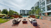 Located at 1348 Passenger St in Chattanooga's vibrant Southside district, this thoughtfully designed outdoor amenity area combines modern resort-style living with distinctive local character. At its centerpiece sits a generously sized swimming pool with sparkling turquoise water, framed by a wide concrete sundeck outfitted with rows of sleek lounge chairs and shade umbrellas in coordinating blue and yellow tones. Surrounding the pool, lush green turf lawns, ornamental grasses, flowering plantings, and mature trees create a verdant, park-like atmosphere that softens the multi-story residential buildings enclosing the courtyard on multiple sides. A standout feature is the repurposed vintage trolley car, transformed into an open-air event and gathering space complete with a raised wood deck, string lights overhead, and bold yellow seating — a nod to Chattanooga's rich transit heritage. Along one perimeter, a striking red brick accent wall is fitted with a series of sculptural water sconces that cascade into a shallow channel below, adding an elegant and tranquil element to the landscape. The mixed-material architecture blends gray siding, brick accents, and large windows, giving the courtyard an upscale yet approachable character ideal for relaxation and socializing.