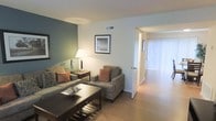 This two-floor space presents a welcoming and well-organized living environment with a clean, contemporary aesthetic. The living area is defined by light hardwood flooring that extends throughout, contributing to a bright and cohesive atmosphere. A teal-painted accent wall adds a subtle pop of color, contrasting tastefully against the neutral white walls that dominate the space.

The living room is furnished with a plush sofa and matching armchair in warm gray tones, accented by rust-colored throw pillows that introduce a touch of warmth. A dark wood coffee table anchors the seating arrangement, while a side table with a classic table lamp provides ambient lighting. A framed artwork piece adorns the accent wall, adding a decorative focal point.

Natural light filters into the adjacent area through vertical blinds, suggesting access to an outdoor-facing window or sliding door. The open sightline between the living room and the adjoining space enhances the sense of flow and openness throughout the floor. Overall, the space balances functional design with comfortable, inviting décor across its two levels.