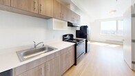 Located at 409 S 23rd St in Tacoma, Washington, this efficiently designed unit offers a thoughtful single-floor layout comprising a combined living and kitchen area, plus a full bathroom. The living space is bright and open, with light wood-tone flooring and crisp white walls that reflect natural sunlight streaming through the windows, creating an airy, clean atmosphere.

The kitchen is well-appointed with modern warm wood cabinetry featuring sleek bar-pull hardware, light-toned countertops, and a stainless steel sink. Full appliances include a black electric range with an overhead exhaust hood, a dishwasher, and a top-freezer refrigerator in a dark finish. The cabinetry extends to the upper walls, maximizing storage capacity. A vivid blue entry door adds a distinctive pop of color and character to the kitchen entryway.

The bathroom is finished in a clean, minimalist style with the same light wood flooring carried throughout the unit. It includes a full soaking tub and shower combination with chrome fixtures, a toilet, and a vanity with a wood-toned cabinet base and light countertop surface. The all-white walls and fixtures contribute to a bright, functional feel. Overall, this unit delivers a modern, cohesive aesthetic well-suited for efficient urban living in Tacoma.