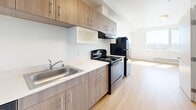 Located at 409 S 23rd St in Tacoma, Washington, this efficiently designed unit offers a thoughtful single-floor layout comprising a combined living and kitchen area, plus a full bathroom. The living space is bright and open, with light wood-tone flooring and crisp white walls that reflect natural sunlight streaming through the windows, creating an airy, clean atmosphere.

The kitchen is well-appointed with modern warm wood cabinetry featuring sleek bar-pull hardware, light-toned countertops, and a stainless steel sink. Full appliances include a black electric range with an overhead exhaust hood, a dishwasher, and a top-freezer refrigerator in a dark finish. The cabinetry extends to the upper walls, maximizing storage capacity. A vivid blue entry door adds a distinctive pop of color and character to the kitchen entryway.

The bathroom is finished in a clean, minimalist style with the same light wood flooring carried throughout the unit. It includes a full soaking tub and shower combination with chrome fixtures, a toilet, and a vanity with a wood-toned cabinet base and light countertop surface. The all-white walls and fixtures contribute to a bright, functional feel. Overall, this unit delivers a modern, cohesive aesthetic well-suited for efficient urban living in Tacoma.