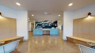 Located at 934 Shorepoint Ct in Alameda, CA, this single-floor leasing office for South Shore Apartments delivers a polished, coastal-modern aesthetic throughout. The centerpiece of the space is a spacious reception area anchored by two curved front desks with distinctive blue geometric tile facades, set against a dramatic large-format wall mural depicting rolling ocean waves. Pendant lighting and recessed fixtures illuminate the area, while polished concrete floors extend throughout, lending an open, contemporary feel. Adjacent to the reception area, a comfortable lounge nook features warm wood-paneled walls, built-in bench seating, woven accent chairs, and wall-mounted sconce lighting. A floor-to-ceiling glazed entry and oversized windows bathe this area in natural light and provide views of the surrounding outdoor landscape, with lush potted palms adding a welcoming, tropical touch. A corridor lined with black-framed glass-panel doors leads to a series of private meeting or office rooms, each offering transparency and access to natural light while maintaining a clean, professional atmosphere. The overall design successfully balances functionality with a refined coastal sensibility, creating an inviting and informative environment for prospective residents.