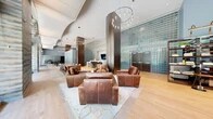 This generously proportioned amenity lounge offers a sophisticated, multi-functional space designed for both relaxation and social engagement. The single-floor layout encompasses a variety of distinct zones that flow seamlessly into one another, unified by warm hardwood flooring, polished stone tile, and soaring arched ceilings that lend an impressive sense of volume throughout.

At the heart of the space, a striking lounge area features rich leather sofas and armchairs arranged around a light-toned area rug, anchored by a dramatic cylindrical column and an eye-catching circular pendant chandelier overhead. A prominent glass block accent wall in cool teal tones serves as a bold architectural focal point, complemented by elegant dark wood double doors.

A recreation area includes a sleek, contemporary billiards table set beneath high ceilings, while a dedicated gaming station adds an entertainment dimension to the space. The library-style lounge zone showcases freestanding dark wood bookshelves and comfortable seating, bathed in natural light pouring through expansive floor-to-ceiling windows framed by textured stone walls.

A communal dining and co-working area features a live-edge wood conference table surrounded by modern chairs, alongside bar-height seating and a built-in bar with a mounted flat-screen television. Throughout, modern pendant lighting and abundant greenery-filled outdoor views enhance the bright, welcoming ambiance.