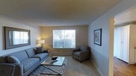 This single-floor residential space offers a thoughtfully arranged living area designed for both comfort and functionality. The living room serves as the central gathering space, furnished with a plush gray sofa and a matching armchair arranged around a sleek glass-top coffee table, all anchored by a patterned area rug. A large window draws in natural light, offering a pleasant view of the surrounding greenery and creating a bright, airy atmosphere during the day. Warm accent lighting from a table lamp adds a welcoming glow to the room during evening hours. The walls feature tasteful framed artwork, contributing to the space's refined yet approachable character. A baseboard heating system runs along the base of the exterior wall, ensuring year-round comfort. The textured ceiling and neutral-toned walls and carpet create a cohesive, understated palette throughout, making the space easy to personalize. Overall, this residence balances practical living with an inviting aesthetic, ideal for those seeking a comfortable and well-appointed home environment.