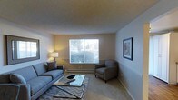 This single-floor residential space offers a thoughtfully arranged living area designed for both comfort and functionality. The living room serves as the central gathering space, furnished with a plush gray sofa and a matching armchair arranged around a sleek glass-top coffee table, all anchored by a patterned area rug. A large window draws in natural light, offering a pleasant view of the surrounding greenery and creating a bright, airy atmosphere during the day. Warm accent lighting from a table lamp adds a welcoming glow to the room during evening hours. The walls feature tasteful framed artwork, contributing to the space's refined yet approachable character. A baseboard heating system runs along the base of the exterior wall, ensuring year-round comfort. The textured ceiling and neutral-toned walls and carpet create a cohesive, understated palette throughout, making the space easy to personalize. Overall, this residence balances practical living with an inviting aesthetic, ideal for those seeking a comfortable and well-appointed home environment.
