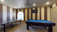 This single-floor space is designed as a dedicated game and recreation room, offering a variety of entertainment options. The room's most prominent features include a full-sized pool table with a blue felt surface and a ping pong table, making it well-suited for social gatherings and leisure activities. A wall-mounted flat-screen TV is positioned on the far wall, adding a media element to the space.

The interior design is bold and distinctive, with alternating brown and cream vertical stripes adorning the primary accent wall, framed by decorative pilasters that lend a structured, theatrical quality to the room. The flooring features a leopard-print carpet that reinforces the room's lively, entertainment-focused aesthetic. Crown molding runs along the upper edges of the walls, adding a touch of architectural refinement.

Lighting is provided by recessed ceiling fixtures and a ceiling fan, ensuring the space remains well-lit and comfortable during use. A arched window with horizontal blinds allows natural light to filter in. The textured ceiling completes the room's layered design, contributing to its overall energetic and inviting ambiance.