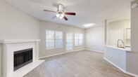 This single-story space is designed with a clean, neutral aesthetic throughout, making it a versatile and functional layout. The main living area serves as the focal point of the space, featuring light wood-look flooring, soft neutral-toned walls, and a white-mantled fireplace that adds warmth and character. A ceiling fan with integrated lighting occupies the center of the textured ceiling, while multiple windows fitted with horizontal blinds allow generous natural light to fill the room. A glass-paneled door provides additional light and access to the exterior.

The kitchen is efficiently arranged in a galley-style layout and features white cabinetry, stainless steel appliances including a refrigerator and range, and a peninsula counter with a built-in sink. This pass-through design connects the kitchen to the living area, enhancing the open feel of the space.

The bedroom continues the consistent flooring and neutral color palette, offering a clean and adaptable space with a ceiling fan overhead. The bathroom is straightforward and functional, featuring a full-sized soaking tub with shower combination, standard fixtures in brushed nickel, and wood-look flooring that ties cohesively to the rest of the unit.