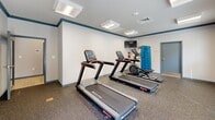 Located at 2001 Amber Leaf Place in Waldorf, Maryland, this fitness center is a dedicated exercise facility designed to provide residents with convenient access to cardiovascular and general workout equipment. The room is finished with light gray walls accented by a navy blue crown molding detail, giving the space a clean, polished appearance. Durable speckled gym flooring covers the entire area, providing a practical and resilient surface suited for high-traffic use. Overhead, flush-mounted rectangular light fixtures provide consistent, even illumination throughout the room, complemented by natural light streaming in through multiple windows along one wall. The fitness center is equipped with multiple Star Trac treadmills featuring integrated digital consoles, an elliptical cross-trainer, and a wall-mounted exercise mat station displaying a stretching guide — offering a well-rounded set of tools for both cardio and flexibility training. A wall-mounted flat-screen television is positioned for easy viewing during workouts. Safety features include a fire extinguisher and smoke detector. The space also benefits from a dedicated entry door and clear fitness center signage, reinforcing its purpose as a structured, community amenity. Overall, the room maintains a tidy, functional atmosphere well-suited for everyday fitness routines.