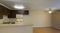 This single-floor residential space offers a functional and straightforward layout centered around an open-concept kitchen and living area. The kitchen is equipped with dark espresso-toned upper cabinetry that provides ample storage, complemented by a light-toned countertop that extends into a breakfast bar-style peninsula — an ideal spot for casual dining or meal prep. White appliances, including a gas range and over-the-counter microwave, are neatly integrated into the kitchen space. The textured ceiling adds a classic residential character throughout, while flush-mounted ceiling lighting ensures the kitchen area remains well-lit. The adjoining living area transitions seamlessly from the kitchen, featuring warm wood-style laminate flooring that adds a welcoming, cohesive feel to the space. Neutral-toned walls throughout provide a clean, versatile backdrop suitable for a variety of interior styles. A wall-mounted split unit offers climate control for year-round comfort. The overall layout maximizes the available footprint, making efficient use of both the kitchen and living zones in a connected, easy-to-navigate single-level design.