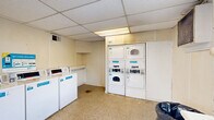 Located at 710 Lone Oak Rd in Paducah, Kentucky, this dedicated laundry facility offers a straightforward and functional space designed to serve shared laundry needs. The room features a drop ceiling with integrated fluorescent lighting that provides even, practical illumination throughout. The walls are finished in a neutral cream tone, and the floor is covered in durable vinyl tile, lending the space a utilitarian character.

The laundry area is equipped with multiple top-load washing machines and stacked dryer units, all branded with a cashless payment system — signage clearly indicates that no coins are required, allowing for modern card or app-based payment. The machines are arranged efficiently along the walls, maximizing the open floor space at the center of the room.

A built-in counter runs along one wall, providing a convenient surface for folding or staging laundry. A window on the side wall allows for natural light and ventilation, complemented by a wall-mounted ventilation unit. The room is accessible through a six-panel exterior door that also brings in additional natural light. An HVAC vent is positioned near the ceiling to maintain comfortable air circulation. Overall, the space is compact yet well-organized, offering essential laundry amenities in a no-frills, utilitarian environment.