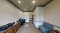 This single-floor office space offers a clean, professional environment designed for both productivity and comfort. The main area features a built-in dark wood workstation with ample desk surface and integrated cabinetry below, providing generous storage and workspace. A computer setup is positioned at the desk, making it ready for immediate use. Adjacent to the workstation, a teal upholstered sofa bench offers a welcoming seating option, ideal for brief consultations or waiting. The space is finished with elegant crown molding that runs along the upper perimeter of the walls, lending a polished, traditional character to the interior. Recessed lighting is installed throughout the ceiling, ensuring even, well-distributed illumination across the room. The walls are painted in a neutral light gray tone, complemented by a subtle chair rail detail that adds architectural interest. Plush, patterned carpeting covers the floor, contributing to a quiet and comfortable atmosphere. An open doorway leads to an adjacent area, suggesting connectivity to further rooms or a hallway beyond. The overall ambiance is professional yet approachable, making this space well-suited for administrative or reception-oriented functions.