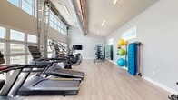 Located at 606 SW 29th Ave in Cape Coral, Florida, this fitness center offers a bright, modern exercise space designed to support a full range of workout routines. The room is defined by soaring vaulted ceilings that lend an open, airy atmosphere, complemented by exposed industrial-style HVAC ductwork that adds a contemporary architectural character. Light wood-look flooring runs throughout, contributing to a clean and welcoming environment.

A striking floor-to-ceiling garage-style window wall anchors one side of the room, flooding the space with abundant natural light and offering direct views of an outdoor pool area and palm trees — creating an energizing backdrop for cardio workouts. A row of treadmills, ellipticals, and a stair climber are positioned along this window wall to take full advantage of the view.

The fitness center is comprehensively outfitted with a cable strength training machine, an adjustable weight bench, a rack of dumbbells, and wall-mounted storage for exercise balls and resistance bands in various sizes. A wall-mounted flat-screen television provides entertainment during workouts, while a large mirror panel enhances the sense of space. Additional windows on the side walls bring in further natural light. A water fountain and exit door are conveniently located at the rear of the room.