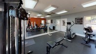 This fitness center, managed by Milestone Management, is a thoughtfully designed workout space that offers a comprehensive range of equipment for both cardio and strength training. The room is finished with cool grey walls accented by a bold orange feature wall, creating an energizing atmosphere that motivates activity. Durable rubber flooring covers the entire space, providing traction and impact absorption throughout.

The cardio section includes a treadmill, an elliptical trainer, and a stationary bike, offering a variety of low- and high-impact workout options. For strength training, the facility features an Inspire functional trainer cable machine, a multi-station weight machine, a leg press, and additional resistance equipment, complemented by a weight bench for free weight exercises. A wall-spanning mirror runs along one side of the room, visually expanding the space and allowing users to monitor their form.

Natural light filters in through multiple windows, keeping the environment bright and open, while flush-mount overhead lighting ensures consistent illumination throughout the day. A wall-mounted television adds an entertainment option during workouts. The fitness center rules are posted on the wall near the entrance, and access is provided through a keyed exterior door, ensuring a secure and organized environment for all users.
