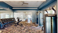 Located at 71 Palomar Avenue in San Luis Obispo, CA, this well-appointed two-story facility offers a thoughtfully designed layout across two generously sized floors. The ground floor serves as the activity hub, anchored by a fully equipped fitness center dressed in bold blue walls, multi-colored carpet tile flooring, and floor-to-ceiling glass partitions. The gym features Matrix-brand cardio and strength equipment, a dumbbell rack, and a ceiling fan, all bathed in natural light from large windows with outdoor views of surrounding greenery. An adjacent kitchenette with white cabinetry and a beverage cooler adds everyday convenience. Classic crown molding and clean white trim carry throughout, lending a refined architectural character to the space. A welcoming lounge area showcases wainscoting, warm hardwood floors, and curated artwork, flowing seamlessly into hallways that connect each functional zone. A charming built-in window seat nook with floor-to-ceiling gridded windows offers a bright, relaxing retreat. The dedicated conference room features warm yellow walls, natural light, and seating for a full team, while a private office with bold houndstooth wallpaper and built-in cabinetry provides a focused workspace. A stylishly appointed bathroom with penny-tile flooring, white subway tile, and pendant lighting rounds out the ground level. A staircase leads to the second floor, which provides additional rooms and closet storage.