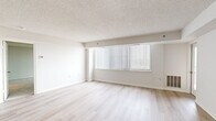 Located at 10301 Grosvenor Pl in Rockville, MD, this two-bedroom, two-bathroom residence offers a well-organized, single-floor layout designed for comfortable living. The open living room is bright and airy, featuring light hardwood flooring, a textured ceiling, and a large window dressed with vertical blinds that allows generous natural light to fill the space. The galley-style kitchen is thoughtfully appointed with granite countertops, warm wood cabinetry, stainless steel appliances including a refrigerator, electric range, built-in microwave, and dishwasher, all set over matching light hardwood floors. Both bathrooms are finished with white tile flooring and granite vanity tops with undermount sinks, maintaining a consistent design language throughout. The primary bedroom is a comfortable, well-sized retreat featuring plush carpeting, a large window with a balcony view of the surrounding landscape, and a closet. The second bedroom also features neutral carpet and ample wall space. A hallway connects the rooms, providing a functional flow through the residence. The clean, neutral palette throughout creates a bright, versatile atmosphere well-suited for a variety of living arrangements.