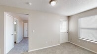 This single-story space offers a practical and straightforward layout designed for comfortable everyday living. The living area is finished with neutral-toned carpet and warm beige walls, complemented by a textured ceiling and a central light fixture. A door with an adjacent window fitted with horizontal blinds allows natural light to filter into the space.

The bedroom features matching carpet flooring, a ceiling fan with a light kit, and a window that brings in additional natural light. A closet door is accessible from within the room, providing convenient storage.

The kitchen is equipped with white upper and lower cabinetry, a speckled stone-look countertop, and a peninsula that separates the cooking area from the adjoining space. Black appliances, including a refrigerator and an electric range with an overhead exhaust, are integrated into the layout. A stacked washer and dryer unit is conveniently located within the kitchen area, adding in-unit laundry functionality.

The bathroom includes wood-look vinyl flooring, a vanity with a stone-look countertop and an undermount sink, a large mirror, and a separate toilet compartment. Towel bars and overhead lighting complete the functional design. Throughout the space, neutral tones and clean finishes create a cohesive and move-in-ready environment.