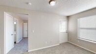 This single-story space offers a practical and straightforward layout designed for comfortable everyday living. The living area is finished with neutral-toned carpet and warm beige walls, complemented by a textured ceiling and a central light fixture. A door with an adjacent window fitted with horizontal blinds allows natural light to filter into the space.

The bedroom features matching carpet flooring, a ceiling fan with a light kit, and a window that brings in additional natural light. A closet door is accessible from within the room, providing convenient storage.

The kitchen is equipped with white upper and lower cabinetry, a speckled stone-look countertop, and a peninsula that separates the cooking area from the adjoining space. Black appliances, including a refrigerator and an electric range with an overhead exhaust, are integrated into the layout. A stacked washer and dryer unit is conveniently located within the kitchen area, adding in-unit laundry functionality.

The bathroom includes wood-look vinyl flooring, a vanity with a stone-look countertop and an undermount sink, a large mirror, and a separate toilet compartment. Towel bars and overhead lighting complete the functional design. Throughout the space, neutral tones and clean finishes create a cohesive and move-in-ready environment.