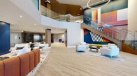 This expansive, single-floor lobby and communal living space makes an immediate impression with its dramatic double-height ceilings and an eye-catching, large-scale geometric mural in vivid blues, oranges, and reds that anchors the main lounge area. Light wood-toned flooring runs throughout, lending warmth and continuity to the open-plan layout. A sleek glass-railed staircase ascends alongside the mural, adding a sculptural architectural element, while circular pendant lighting fixtures overhead contribute a contemporary, refined ambiance.

The main lounge is thoughtfully arranged with multiple seating zones, including plush white armchairs grouped around a low coffee table, and a curved, terracotta-hued sectional sofa positioned beneath a large wall-mounted display screen set against a bold navy blue accent wall. A built-in live moss feature adds a natural, organic touch beneath the staircase.

Glass-partitioned meeting and co-working rooms line the perimeter, offering transparency and accessibility while maintaining acoustic separation. A secondary lounge nook features distinctive violet accent chairs alongside dark marble wall panels, curated artwork, and a service counter, creating a calm, gallery-like retreat. The main entry is framed by black-trimmed glass doors that draw in natural light and provide views to the exterior, reinforcing the space's open, welcoming character throughout.
