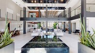 This expansive two-floor space is a masterclass in contemporary design, with the main level commanding the majority of the footprint and an open upper level overlooking the floors below via a sleek metal railing system. At the heart of the main level sits a dramatic double-height atrium, anchored by a striking recessed water feature filled with river stones, surrounded by curated lounge seating and bold structural columns. Tall floor-to-ceiling windows and glass-paneled entry doors flood the space with natural light, offering views to the surrounding exterior. A refined staircase with vertical wood slat detailing connects the two levels, serving as an architectural focal point. The main level also features distinct gathering zones, including a dining-style seating area beneath a large-format stone accent wall, and a separate lounge wing with floor-to-ceiling black-framed windows and wood-slat ceilings. The second floor hosts a social lounge with a full wet bar, designer pendant lighting, a wave-form acoustic ceiling feature, built-in seating with wood surrounds, and warm wide-plank hardwood flooring. Additional areas on this level offer window bench seating and access to outdoor views. Throughout, the design balances industrial and organic materials — concrete, wood, stone, and metal — creating a cohesive, sophisticated ambiance.