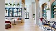 Located at 3712 Stewart Rd in Doraville, Georgia, this beautifully designed lobby space offers a sophisticated and welcoming first impression. The interior is defined by soaring double-height ceilings that flood the open floor plan with natural light through expansive black-framed floor-to-ceiling windows, creating an airy and upscale ambiance throughout.

The main lounge area features light wide-plank hardwood flooring and is furnished with a curated mix of seating — including a neutral-toned sofa accented with colorful pillows, sleek accent chairs in warm wood tones with velvet upholstery, and cane-back chairs paired with marble-topped bistro tables. A bold dark navy built-in shelving unit with arched detailing anchors one wall, displaying decorative vessels and lush trailing plants.

A standout wet bar area is finished in vibrant teal with arched alcoves, mosaic tile backsplash, warm wood cabinetry, and a patterned hexagonal tile floor — providing a striking design focal point. Adjacent to this is a communal workspace with bar-height seating alongside a dark island counter.

A grand staircase with light wood treads and sleek black metal railings connects to an upper mezzanine level. Gold pendant details and hanging greenery add warmth and texture, completing this thoughtfully designed, design-forward communal space.