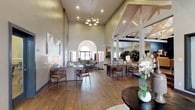 This single-floor space is defined by its dramatic architectural details and open, multi-functional layout. The main lounge area commands attention with soaring vaulted ceilings supported by white-painted exposed beam trusses set against deep navy accent walls. A full-height stacked stone fireplace anchors one end of the room, while generous seating arrangements—including a caramel leather sofa, wingback chairs, and neutral sectional sofas—create an inviting, lounge-like atmosphere. An arched window floods the space with natural light, complementing the warm hardwood-style flooring found throughout.

The entry and reception area features similarly high ceilings, an elegant chandelier, and a welcoming arrangement of seating and work surfaces. Recessed lighting and wall-mounted artwork contribute to a polished, contemporary feel. A hallway lined with additional rooms or offices extends from this central hub.

The kitchen area is equipped with stainless steel appliances, warm honey-toned cabinetry, granite countertops, and a large island with bar seating on multiple sides. Pendant lighting hangs above the island, reinforcing the open and functional design. Throughout, the consistent use of hardwood-style flooring, neutral wall tones, and carefully curated furnishings creates a cohesive, well-appointed environment suited for both social gatherings and everyday use.