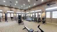 This fitness center offers a comprehensive workout environment designed to accommodate a variety of exercise needs. The space is defined by warm exposed brick walls that lend an industrial yet inviting character, complemented by rows of large windows that line the upper perimeter of the room, flooding the interior with natural light. Recessed lighting and ceiling fans ensure the space remains bright and well-ventilated throughout.

The floor is finished with durable striped carpeting suited for high-traffic athletic use. The layout is thoughtfully organized, with a generous selection of cardio equipment — including treadmills, elliptical trainers, stair climbers, and stationary bikes — arranged in orderly rows. Strength and resistance machines are positioned along the sides, providing users with a full range of training options without overcrowding the floor space.

The room's generous proportions allow for comfortable movement between stations, making it practical for simultaneous use by multiple individuals. The combination of rustic brick detailing, abundant natural light, and modern fitness equipment creates a motivating and functional atmosphere, making this fitness center a standout amenity well-suited for daily use.