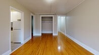 This well-organized one-bedroom, one-bathroom unit offers a practical and classic layout across a single floor. The living room is the largest area of the home, characterized by warm, polished hardwood floors that extend throughout the space, neutral gray walls, and multiple windows that invite natural light. A white cast-iron radiator provides traditional heating, complementing the building's classic character. A hallway leads to the cozy bedroom, which features the same gleaming hardwood floors, a window with views of the surrounding urban streetscape, and a dedicated radiator. An open closet with a hanging rod provides convenient storage. The kitchen is efficiently designed with white cabinetry, a tile backsplash, dark laminate countertops, a stainless steel sink, and a white electric range. A window above the counter and a radiator keep the space bright and warm. The bathroom is fully tiled in soft gray, featuring a wall-mounted sink, toilet, bathtub, and a white radiator, all accented by a natural light window. Two hallways connect the rooms, ensuring smooth circulation throughout. The unit's consistent use of hardwood flooring and neutral tones creates a cohesive, understated aesthetic throughout.