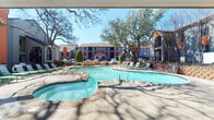 This outdoor cabana structure serves as a dedicated poolside retreat within a residential community. The single-level structure is defined by a steeply pitched, wood-paneled ceiling that lends warmth and architectural character to the open-air space. The deck is constructed from natural wood planking and opens broadly toward the community's freeform swimming pool, which features a connected spa and a spacious stone-paved surround dotted with lounge chairs.

The covered seating area is furnished with wicker lounge chairs and sofas fitted with green cushions, arranged around a low coffee table atop turf-style area rugs. A wall-mounted flat-screen television is affixed to the corrugated metal accent wall at one end, creating a comfortable outdoor viewing area. Decorative elements including potted plants, sculptural vessels, and a console table complete the interior styling.

The cabana opens fully on one side, framing unobstructed views of the pool and the mature trees that shade the surrounding deck. The outdoor setting is bright and well-lit by natural sunlight, contributing to a relaxed, resort-like atmosphere. The structure is integrated into a larger multi-building community, with balconied residential buildings visible in the background.