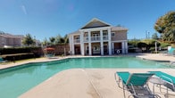 This outdoor amenity space is centered around a generously sized swimming pool with clear, blue-green water, surrounded by a spacious concrete pool deck. The deck is furnished with teal lounge chairs and chaise lounges, providing ample space for sunbathing and relaxation. Vibrant red market umbrellas offer shaded seating areas with patio tables and chairs, ideal for outdoor dining and socializing.

A covered patio area, distinguished by classic white columns and a pitched roof, anchors the space architecturally. Beneath this structure sits a well-equipped outdoor kitchen station, featuring a built-in stainless steel grill set into a stacked stone surround with storage drawers below — a standout feature for outdoor entertaining.

The pool area is enclosed by black wrought iron fencing, with lush landscaping including manicured hedges and mature trees providing a natural backdrop. Views of the surrounding residential buildings are visible beyond the greenery, adding context to the community setting. The overall ambiance is bright, open, and inviting, benefiting from abundant natural light and a well-maintained outdoor environment suited for year-round leisure activities.