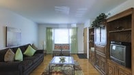 This single-floor space offers a warm and inviting atmosphere anchored by a well-appointed living area. The room features rich hardwood flooring that extends throughout, lending a sense of warmth and continuity to the space. Natural light flows in generously through a large window dressed with elegant green curtains, creating a bright and airy feel. A pair of accent chairs with decorative pillows are positioned near the window, complemented by a plush sofa with coordinating cushions, making the seating arrangement both functional and inviting. A glass-top coffee table rests atop a richly patterned area rug, adding a refined focal point to the room. A substantial dark wood entertainment unit anchors one side of the space, offering ample display and storage capability with its combination of shelving, cabinetry, and drawers. Trailing greenery atop the unit adds a natural, organic touch to the interior. The smooth, light-toned ceiling with recessed lighting panels contributes to the clean and uncluttered aesthetic of the room. Overall, the space balances classic furnishings with practical design, creating a comfortable and well-organized environment suited for everyday living.
