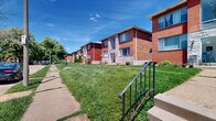 This expansive residential complex comprises a row of two-story buildings that collectively span an impressive total floor area, offering a significant footprint along a quiet, tree-lined street. The buildings showcase a classic mid-century architectural style, constructed primarily with red and tan brick exteriors, accented by stone facade panels that add visual texture and character. Large windows are featured throughout, allowing for abundant natural light across all units.

Each building entrance is approached via concrete steps with decorative wrought iron railings, lending a traditional charm to the streetscape. The grounds between the buildings and the sidewalk are covered with neatly maintained green lawns, with mature trees and small plantings providing shade and greenery. A continuous sidewalk runs the length of the complex, connecting each entrance and creating a cohesive, walkable environment.

From the street, the buildings present a uniform yet visually engaging profile, with subtle variations in facade materials breaking up the repetition. The surrounding neighborhood is well-established, with tree canopy and open street views contributing to a pleasant outdoor ambiance. The scale of the complex, with multiple structures extending along the block, reflects a well-organized and sizeable residential development suited for a broad range of occupants.