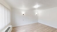 Located at 310 E Foothill Blvd in Pomona, California, this efficient single-floor unit offers a well-organized layout comprising a living area and a full bathroom. The living space is finished with light-toned wood laminate flooring that runs throughout, creating a clean and cohesive aesthetic. Crisp white walls and a textured ceiling contribute to a bright, open atmosphere, while stylish wall-mounted sconces provide warm accent lighting alongside a centrally positioned ceiling fixture. A window fitted with vertical blinds allows natural light to filter in and provides a view of the exterior, while a wall-mounted HVAC unit ensures year-round climate control. A full-size refrigerator is conveniently positioned near the entry door, adding practical functionality to the space. The bathroom features a full bathtub and shower combination with smooth, light-colored surround panels, complemented by a vanity mirror with built-in shelving. The overall design is clean, neutral, and move-in ready, making it a functional and straightforward space suited for everyday living.