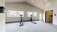 Located at 1641 Belsly Blvd in Moorhead, Minnesota, this fitness center offers a dedicated exercise space designed to support a well-rounded workout routine. The room features a striking vaulted ceiling that lends an open, airy feel, complemented by recessed lighting that keeps the space bright and functional. A row of large windows along one wall floods the interior with natural light while providing views of the surrounding community. The flooring consists of neutral, patterned carpet tiles that provide a comfortable underfoot surface throughout the space. The exercise room is equipped with multiple cardio machines, including treadmills and elliptical trainers, arranged to allow ample room for movement. A dedicated strength training area includes a dumbbell rack and additional fitness equipment, supported by wall-mounted mirrors that help users monitor their form. An accent wall finished in deep navy tile adds a modern, polished aesthetic contrast to the otherwise clean, neutral palette of white and light gray walls. The facility also includes access to a restroom, adding convenience for users. The overall design is clean, contemporary, and purposeful, creating an inviting environment well-suited for daily fitness routines.