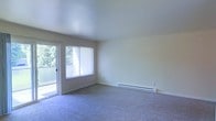 This single-floor space features a spacious living area finished with soft, wall-to-wall carpeting in a neutral blue-grey tone that adds warmth underfoot. The room is bathed in natural light, courtesy of a large sliding glass door that opens to an outdoor balcony or patio area, offering views of mature greenery beyond. Vertical blinds provide adjustable light control and privacy. The smooth, painted walls in a clean off-white tone complement the bright, airy atmosphere of the room. A baseboard heater runs along one wall, providing an efficient and unobtrusive heating solution. The ceiling is smooth and generously high, contributing to the open, comfortable feel of the space. The layout is open and versatile, offering ample room for a variety of furniture arrangements. Overall, the space presents a clean, functional, and well-lit environment with a calm, neutral aesthetic throughout.