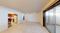 This single-floor space offers a straightforward and functional layout designed for comfortable everyday living. The main living area is generously sized, featuring light-toned wall-to-wall carpeting that adds warmth underfoot. A textured ceiling runs throughout, and multiple ceiling vents ensure consistent climate control across the space. Natural light enters through a dark-framed sliding glass door on one side, which provides direct access to an outdoor area, adding both convenience and openness to the living space.

Adjacent to the main living area, the kitchen is visible through an open doorway and features wood-toned cabinetry and white appliances, including a full-size refrigerator. The kitchen layout appears compact yet functional, with sufficient counter and storage space for daily use. A hallway visible from the living area leads further into the unit, suggesting additional rooms beyond the main living and kitchen zones. The overall color palette — white walls, neutral carpeting, and warm wood accents — creates a clean, understated interior that serves as a blank canvas for personalization.