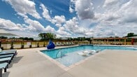 Located on Martin Road SW in Huntsville, Alabama, this two-story residential community is designed with comfort and outdoor living in mind. The property's most striking feature is its expansive resort-style pool area, which serves as a central gathering space for residents. The generously sized swimming pool showcases clear, blue water and includes built-in in-water lounge chairs, perfect for relaxing in the Alabama sun. A dedicated spa with water features adds an additional layer of leisure to the outdoor environment. Surrounding the pool, a broad concrete deck is furnished with multiple chaise loungers and seating areas, offering ample space for sunbathing and socializing. A wood-framed pergola at the perimeter provides shaded seating with picnic-style tables, ideal for outdoor dining or casual gatherings. The pool area is enclosed by a black iron fence, ensuring privacy while maintaining open sightlines to the lush greenery and open skies beyond. The two-story building facades visible from the pool area feature classic architectural detailing with neutral siding, balconies, and brick accents. Landscaping throughout the outdoor space adds color and texture, enhancing the overall ambiance of this well-appointed community.