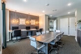 This well-appointed business center and conference room offers a professional and functional environment designed to support a variety of work and meeting needs. The space is anchored by a large rectangular conference table surrounded by sleek, modern chairs, providing ample seating for group meetings or collaborative sessions. Built-in desktop workstations along one wall, illuminated by stylish sputnik pendant lights, offer dedicated computer terminals ideal for individual work tasks. A textured accent wall in a rich dark mosaic pattern serves as a striking focal point, complemented by a wall-mounted flat-screen display and a row of dark cabinetry with granite countertops below. Natural light fills the room through multiple large windows dressed with teal panel drapes, offering views of the surrounding outdoor environment. Framed abstract artwork and decorative sculptural wall elements — including an arrow, "@" symbol, and question mark — add personality and visual interest throughout the space. Plush multi-tone carpeting runs underfoot, adding warmth and acoustic comfort. Storage is further supported by interior doors leading to closet or utility spaces. The overall design balances contemporary aesthetics with practical, productivity-focused amenities.
