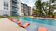 This expansive outdoor amenity area is designed around a generously sized resort-style swimming pool that serves as the central focal point of the space. The pool features built-in water jets, a wading shelf with in-water lounge chairs in a bold coral-orange hue, and a dedicated spa section, creating a layered aquatic experience. Smooth stone coping surrounds the pool deck, which offers ample space for additional seating and lounging throughout.

The multi-story building facade, constructed with a combination of warm wood paneling, brick, and neutral stucco, wraps around the courtyard and provides a striking architectural backdrop. Screened balconies and open-air terraces on the upper levels overlook the pool area below.

A shaded pergola retreat is situated at one end of the courtyard, outfitted with a canvas canopy, decorative planters, hammocks, and casual seating, offering a quiet alternative to the poolside energy. Nearby, a landscaped garden bed enclosed by dark horizontal wood fencing features tropical and drought-tolerant plantings, including yucca and ornamental grasses. Mature oak trees provide natural canopy and shade throughout the courtyard, enhancing the overall ambiance and connecting the space to its natural surroundings.