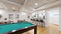 This thoughtfully designed single-floor space seamlessly blends entertainment and everyday living across an open, well-appointed layout. Warm hardwood floors run throughout, unifying each distinct zone while complementing the crisp white walls and beamed ceiling overhead.

The living room is anchored by striking exposed brick columns that frame a wall-mounted flat-screen TV, adding rustic character to the otherwise modern aesthetic. A brick fireplace nook on one side adds architectural interest, while comfortable sectional and swivel lounge seating create an inviting gathering space.

At the heart of the layout, a full-size pool table serves as the centerpiece of a dedicated recreation zone, positioned beneath a coffered ceiling with recessed lighting. Adjacent to it, the dining area features a round table with mid-century modern chairs, accented by framed landscape artwork and a statement wall sconce.

The galley-style kitchen is clean and functional, outfitted with white cabinetry, gold hardware, a subway tile backsplash, quartz countertops, and a black matte sink. It opens to the main area through a graceful arched pass-through bar counter lined with upholstered bar stools and illuminated by pendant lights, making it ideal for entertaining. A built-in bookshelf adds a personal touch nearby.