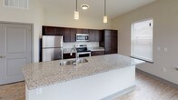 Located at 115 Dayton St in Hamilton, Ohio, this thoughtfully designed single-floor residence offers a clean, modern aesthetic with quality finishes throughout. The open-concept kitchen is a standout feature, boasting rich dark cabinetry, granite countertops, stainless steel appliances, and a large center island with a built-in sink — ideal for cooking and entertaining. Light wood-style flooring runs through the main living areas, complementing the neutral color palette and creating a warm, cohesive atmosphere. The bright living area welcomes natural light through multiple windows fitted with blinds, and a ceiling fan adds both style and comfort. Bedrooms are generously sized, each with plush carpeting and windows that bring in ample natural light. The primary bedroom connects directly to an en-suite bathroom featuring a granite-topped vanity with dark cabinetry and a tiled walk-in shower with glass enclosure. A second full bathroom includes a soaking tub with a tiled surround and wood-look flooring. A dedicated laundry room with wire shelving and washer/dryer hookups adds everyday convenience. Throughout the space, neutral tones, quality materials, and functional layouts combine to create a comfortable and contemporary living environment.
