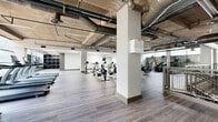 This large-scale fitness facility offers a comprehensive workout environment defined by its industrial-inspired design and generous open floor plan. The space is characterized by exposed concrete ceilings, visible ductwork, and structural columns that lend a raw, urban aesthetic throughout. Light wood-look flooring flows through the cardio zones, while rubber flooring grounds the strength training areas for safety and durability.

The facility is thoughtfully divided into distinct workout zones. A dedicated cardio area accommodates rows of stationary bikes, rowing machines, treadmills, and ellipticals, benefiting from abundant natural light streaming through large floor-to-ceiling windows that offer views of surrounding greenery. Adjacent to this is a spacious strength training zone, equipped with a comprehensive array of selectorized machines, free weights, benches, and a punching bag, set against exposed brick accent walls.

A mezzanine level overlooks the main floor via industrial-style metal mesh railings, connected by an open staircase. Built-in shelving units provide convenient equipment storage. Ceiling fans, suspended linear lighting, and skylights contribute to a well-lit, well-ventilated environment. The facility's multi-zone layout and generous proportions make it well-suited for high-capacity use in a commercial or institutional setting.