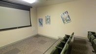 This single-floor screening room is designed to deliver an enjoyable movie-watching experience in a compact, theater-inspired setting. The room is anchored by a large wall-mounted retractable projection screen positioned at the front, paired with overhead lighting that ensures a comfortable viewing environment. Rows of plush, dark green theater-style seats are arranged in a tiered layout, allowing clear sightlines to the screen from every seat. The neutral, light yellow walls provide a clean backdrop and are decorated with colorful animated movie posters, including titles such as Planes, Monsters University, and Tinker Bell, lending the space a distinctly family-friendly and whimsical atmosphere. The flooring features a patterned carpet with a mix of neutral tones, adding warmth and acoustical comfort to the room. Baseboard details and clean wall finishes contribute to a well-maintained, polished look. The overall ambiance is intimate and inviting, making this screening room well-suited for children's movie viewings, family gatherings, or casual group entertainment within a residential or community setting.