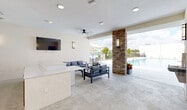 Located at 22555 SW 107th Ave in Miami, FL, this single-story amenity space offers a well-appointed outdoor recreation area designed for community use. The layout seamlessly blends a covered indoor-outdoor lounge with direct access to a large, resort-style swimming pool surrounded by a spacious deck. The covered lounge area features polished concrete flooring, recessed lighting, and ceiling fans that provide comfort in Miami's warm climate. A wall-mounted flat-screen TV and modern sectional seating with neutral-toned cushions create an inviting entertainment atmosphere. A built-in wet bar or service counter with wood-accented paneling anchors one side of the space, adding both functionality and style. A striking stacked-stone column serves as a bold architectural feature, framing the open transition between the covered lounge and the poolside terrace. The pool area is enclosed by clean, cream-colored walls and features lounge chairs and dining furniture set on turf and concrete surfaces under the open sky. Two private bathrooms, accessed through sleek gray doors with wall sconce lighting, provide convenience for guests. The overall design aesthetic is clean and contemporary, with neutral tones, modern fixtures, and thoughtful amenities throughout.